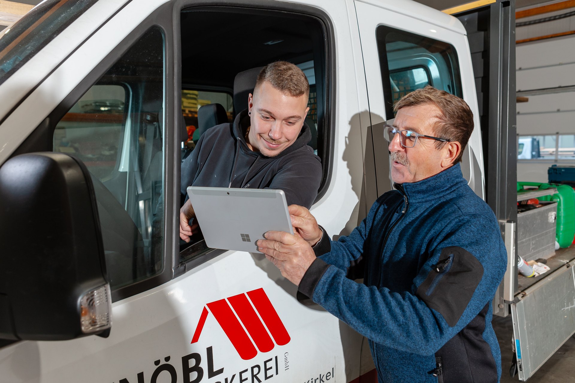 Zwei Mitarbeiter besprechen sich mit einem Tablet am Fenster eines Transporters der T. Knöbl Dachdeckerei