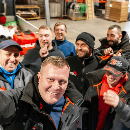 Sieben lächelnde Männer posieren für ein Selfie in einem Lagerhaus, einige tragen schwarze Hilti-Jacken.
