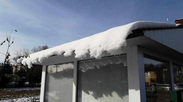 Dicke Schneedecke auf einem weißen Dachvorsprung mit Eiszapfen und großen Fenstern darunter.