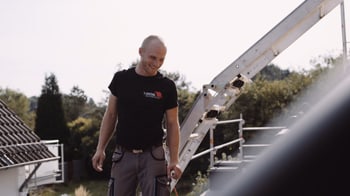 Ein lächelnder Dachdecker in schwarzem T-Shirt mit rotem Logo steht auf einem Dach neben einer Leiter.