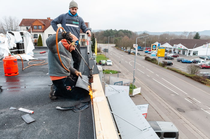 Zwei Dachdecker verlegen Bitumenbahnen mit einem Brenner auf einem Flachdach.