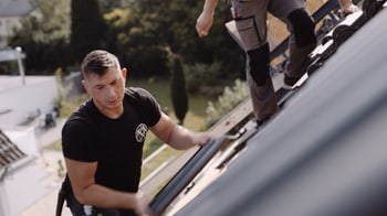Zwei Dachdecker bei der Arbeit auf einem Dach mit dunklen Ziegeln, einer hält einen Ziegelstein in der Hand.