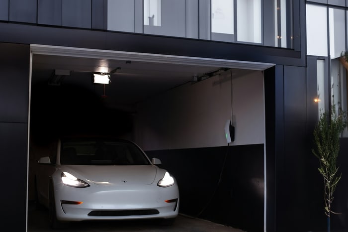Weißes Tesla Model 3 fährt aus einer dunklen Garage mit offenem Garagentor und Ladestation.