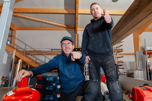 Zwei Handwerker in einer Werkstatt: Der ältere Mann kniet und zeigt, der jüngere steht und gibt Daumen hoch.