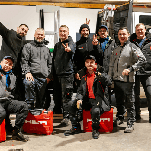 Zehn lächelnde Handwerker in Arbeitskleidung, einige zeigen das Victory-Zeichen, mit roten Hilti-Werkzeugkoffern.