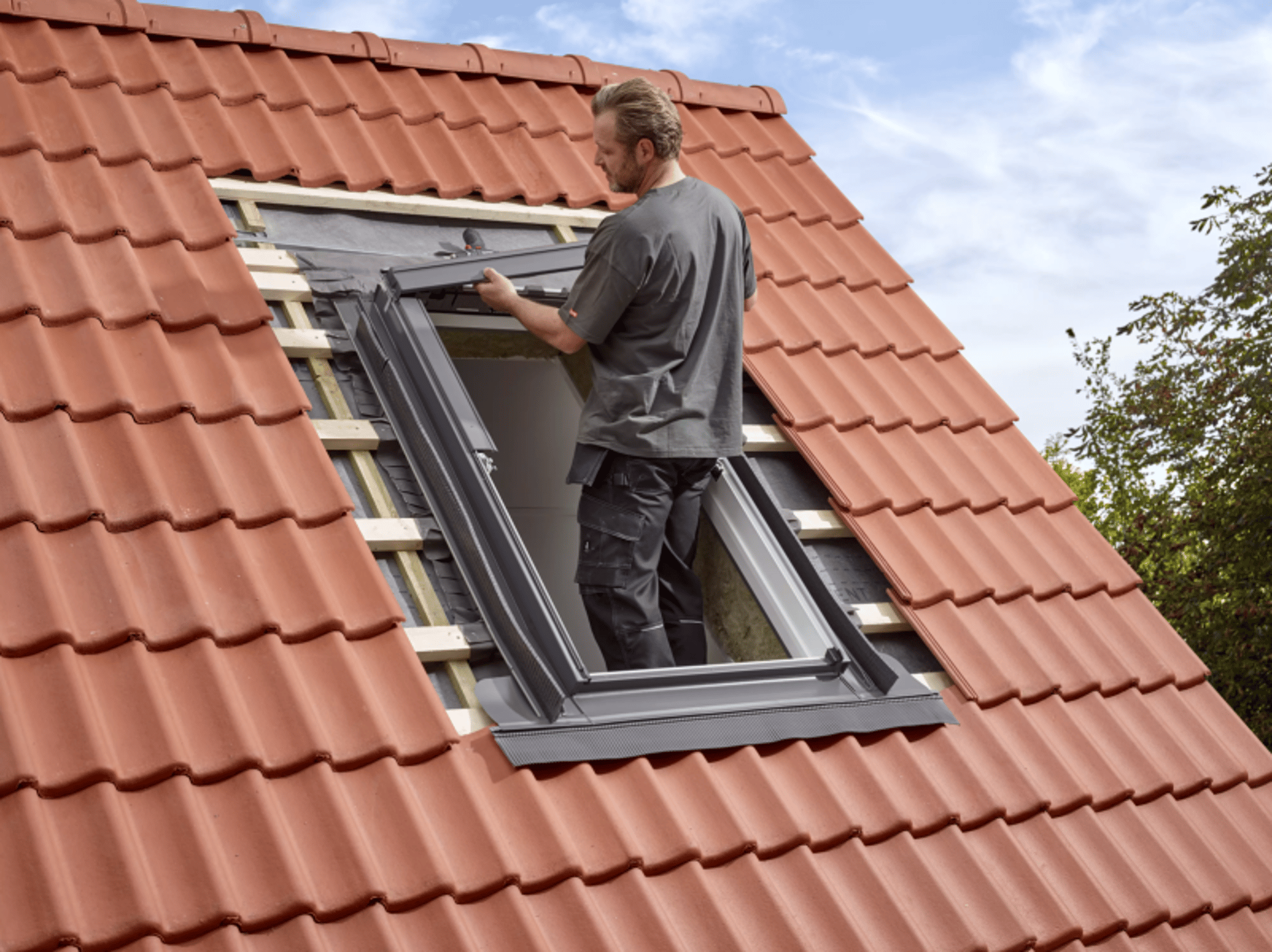 Ein Handwerker installiert ein Dachfenster auf einem Ziegeldach mit roten Dachziegeln.