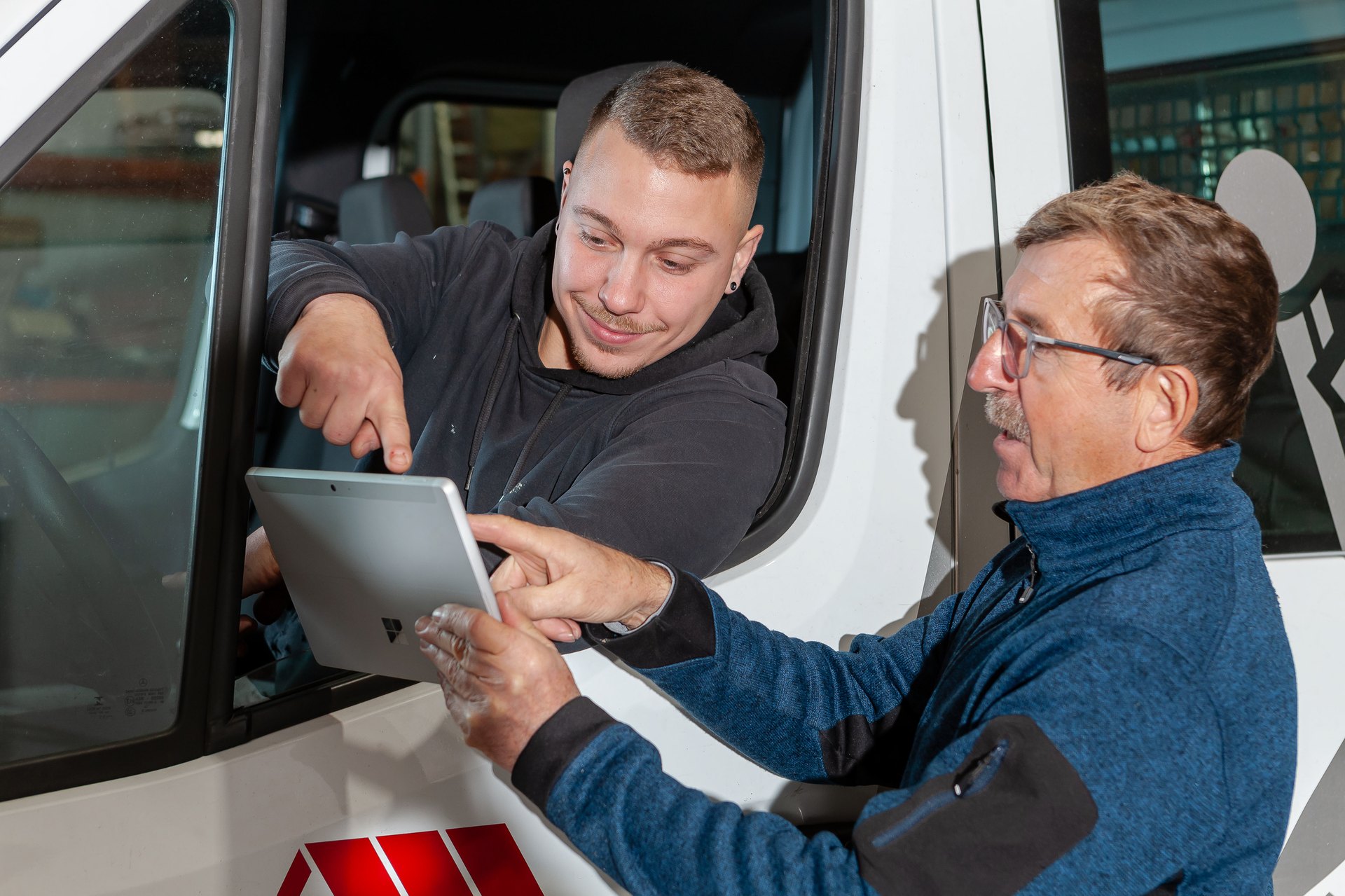 Zwei Männer schauen auf ein Tablet, einer sitzt im Transporter und zeigt auf den Bildschirm.