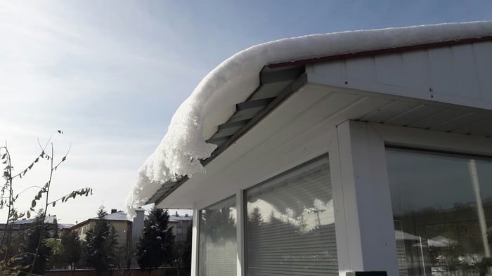 Dicke Schneeschicht auf dem Dach eines weißen Gebäudes mit großen Fenstern unter blauem Himmel.