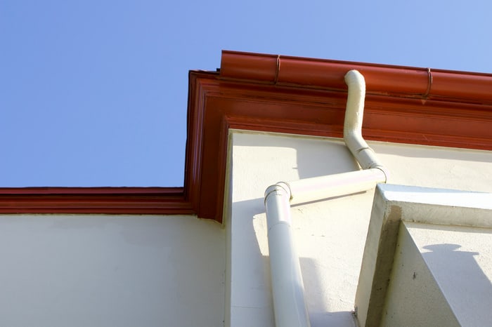 Rote Dachrinnen und weiße Fallrohre an einem Gebäude vor blauem Himmel.