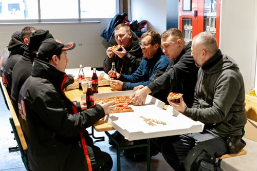 Sieben Männer in Arbeitskleidung essen Pizza und trinken Cola an einem Holztisch.