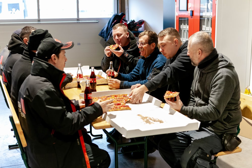 Sieben Männer in Arbeitskleidung essen Pizza und trinken Cola an einem Holztisch.