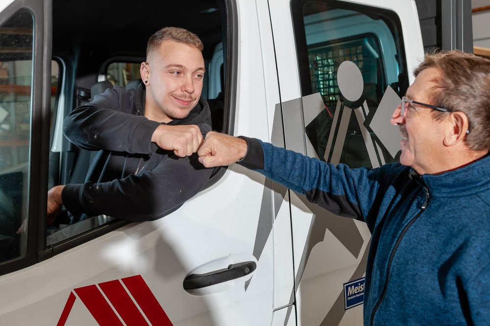 Zwei Männer geben sich einen Fauststoß durch das Fenster eines weißen Lieferwagens.