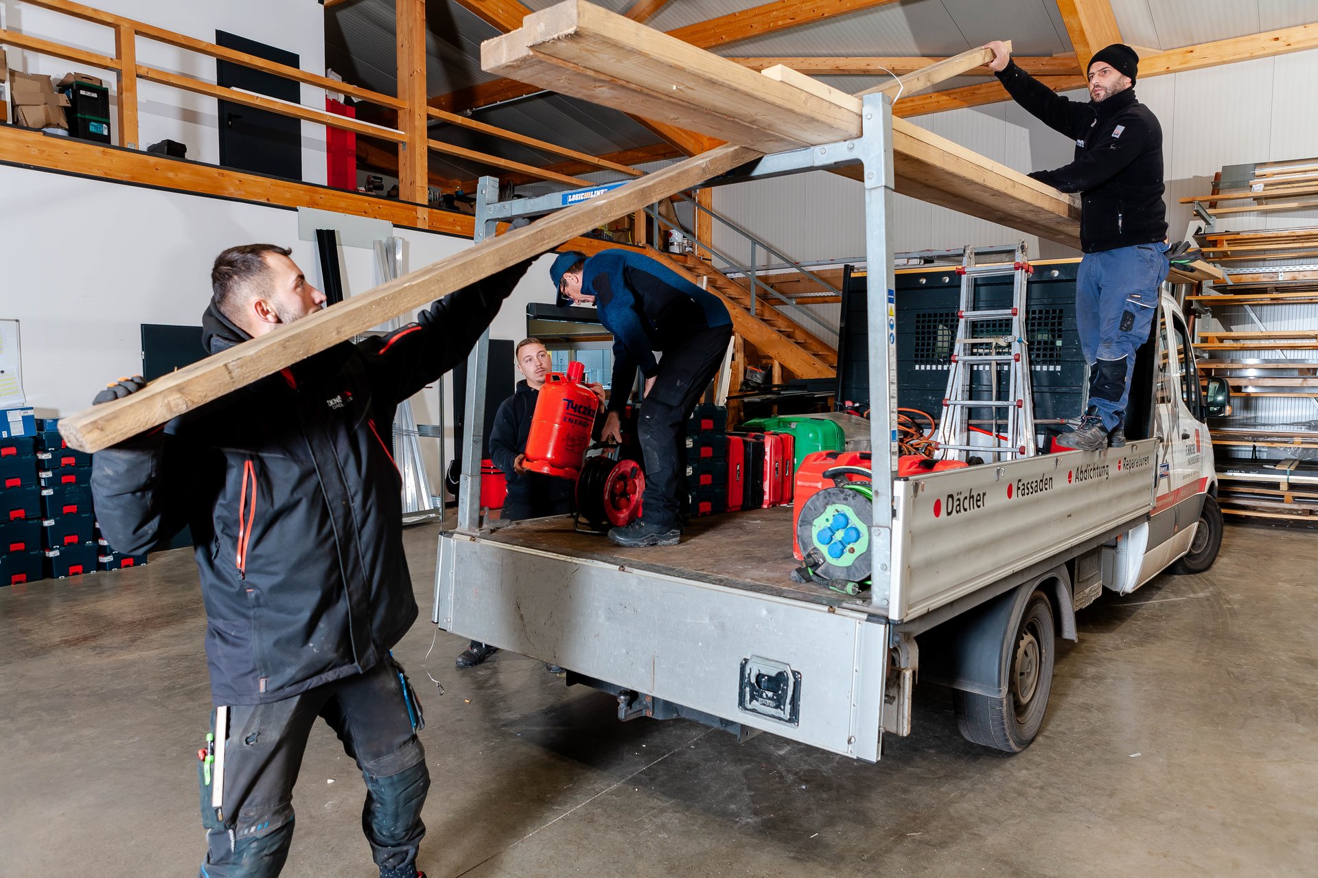 Drei Männer laden Holzbretter und Werkzeuge von einem weißen Lieferwagen in eine Lagerhalle.