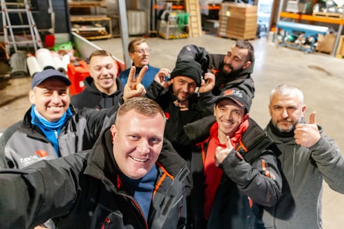 Sieben Männer in Arbeitskleidung posieren lächelnd in einer Lagerhalle, einer zeigt das Friedenszeichen.