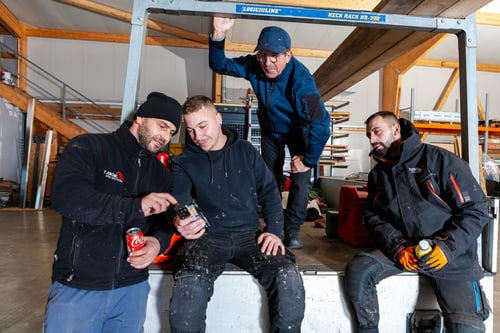 Vier Bauarbeiter machen Pause in einer Lagerhalle, drei sitzen, einer lehnt; eine Cola-Dose ist sichtbar.