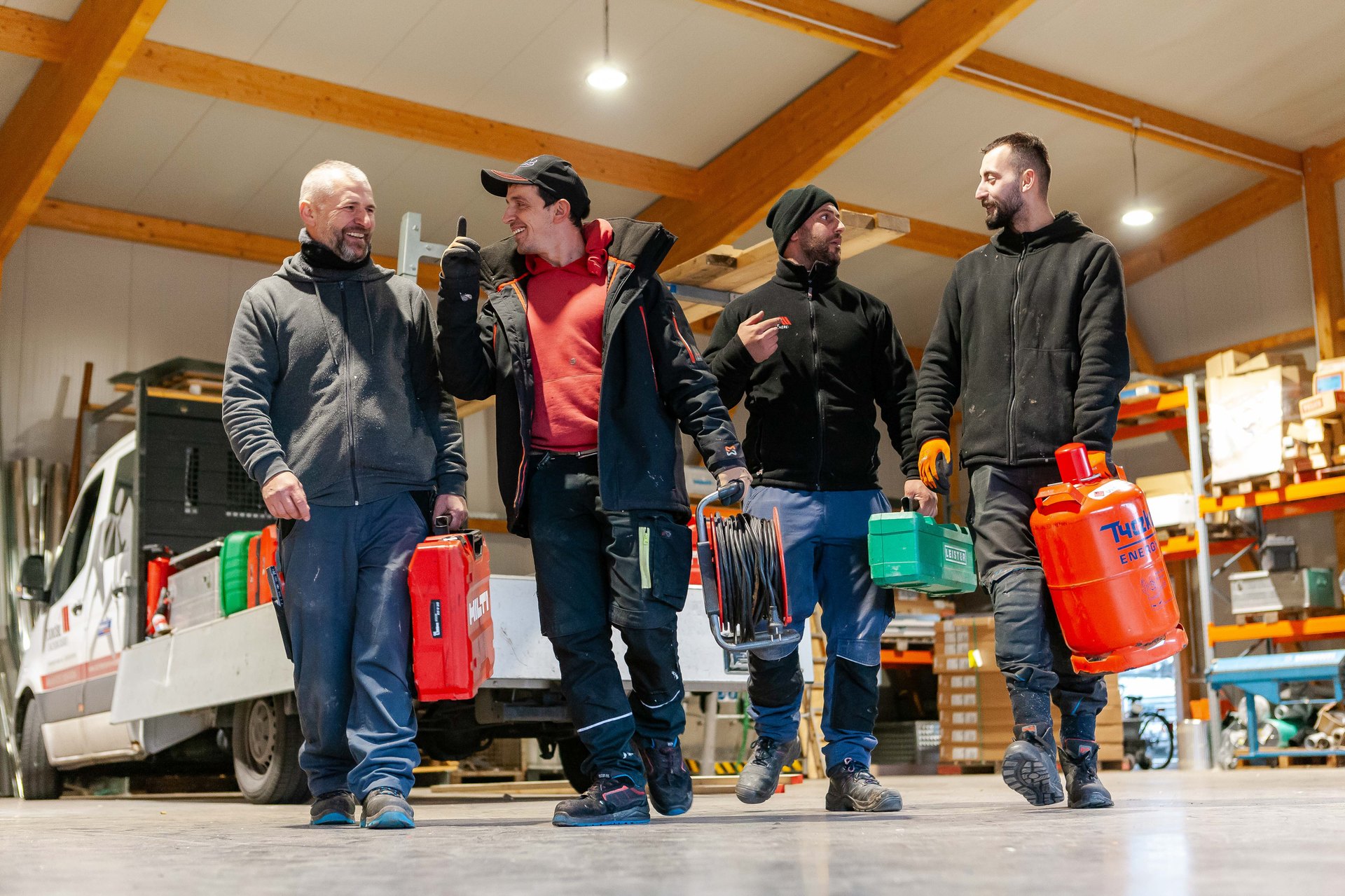 Vier Handwerker tragen Werkzeugkästen und Kanister in einer Werkstatt.